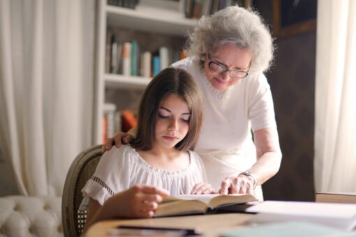 A teenager is reading a book at a desk, with an older woman over her shoulder pointing to something on the page.